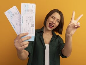 joyeuse-jolie-femme-caucasienne-clignote-yeux-gestes-signe-main-victoire-tenant-billets-avion-orange